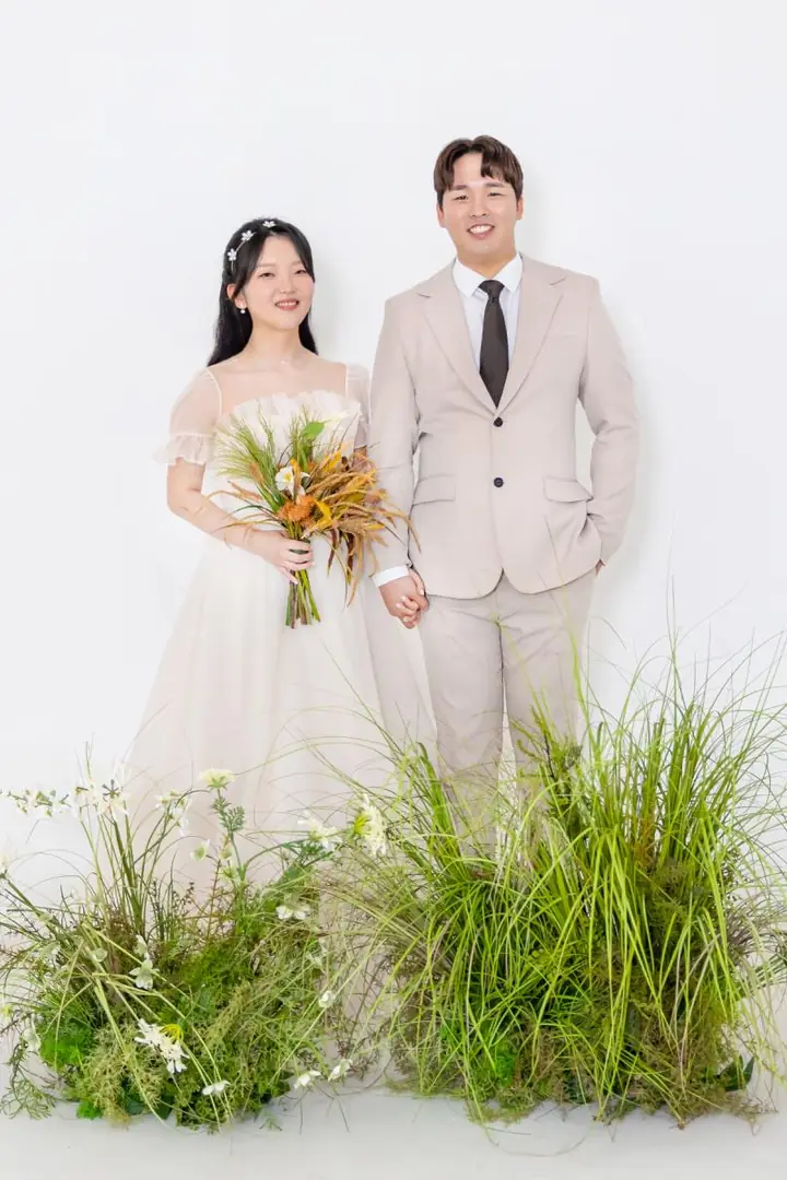 흰 배경 앞 들꽃 사이에 나란히 선 두 사람