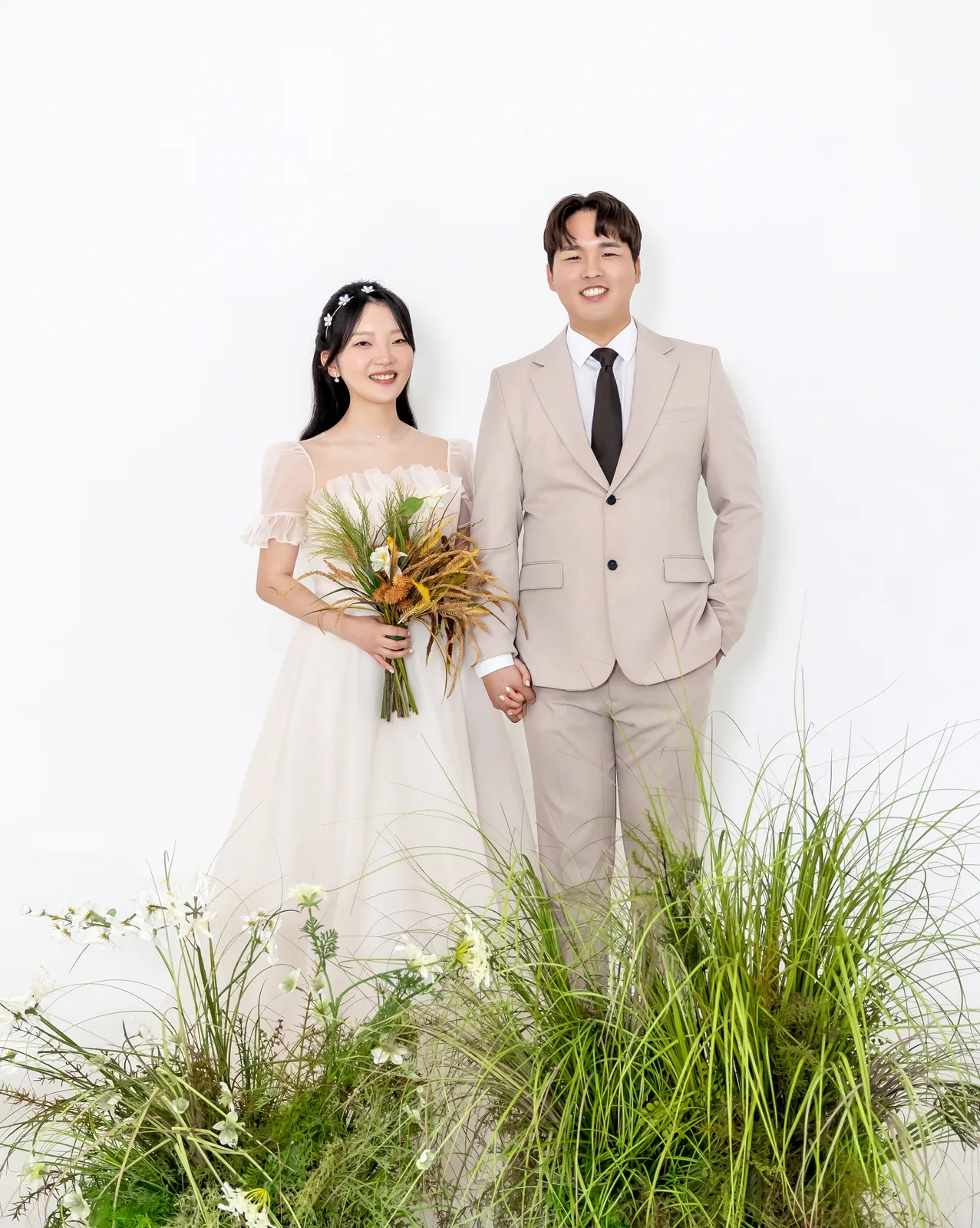 흰 배경 앞 들풀 사이에 나란히 선 두 사람